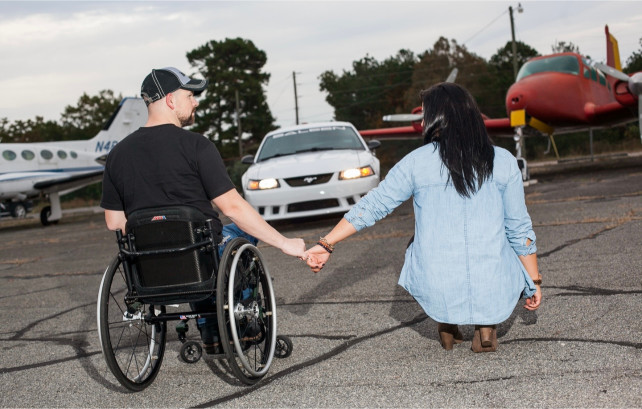 A man in a wheelchair and a woman hold hands while facing an airfield with small planes and a car behind them.