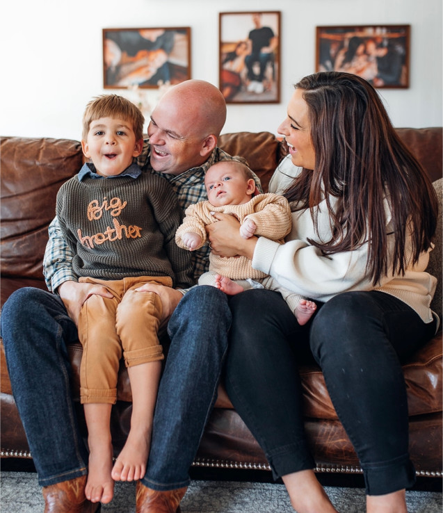 A family of four sits on a brown leather couch. The father holds a boy in a "big brother" sweater, and the mother holds a baby in a beige outfit. Three framed photos hang on the wall behind them.