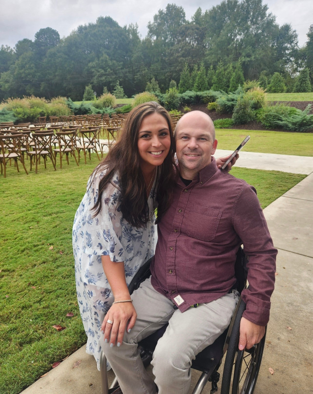 A woman in a floral dress stands next to a man sitting in a wheelchair, both smiling outdoors with empty wooden chairs and trees in the background.