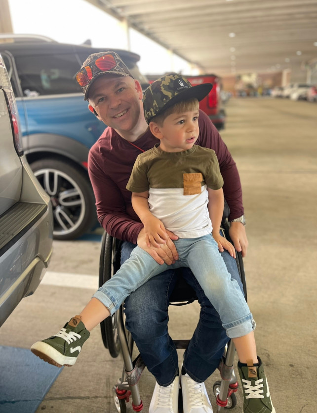 A man in a wheelchair is holding a young boy on his lap in a parking lot. Both are wearing caps and casual clothing.